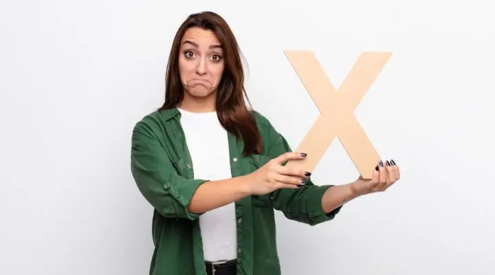 Una mujer con una camisa verde sostiene una gran letra "X" de madera y hace una expresión facial decepcionada o insegura, lo que refleja que está bien equivocarse al tomar decisiones.