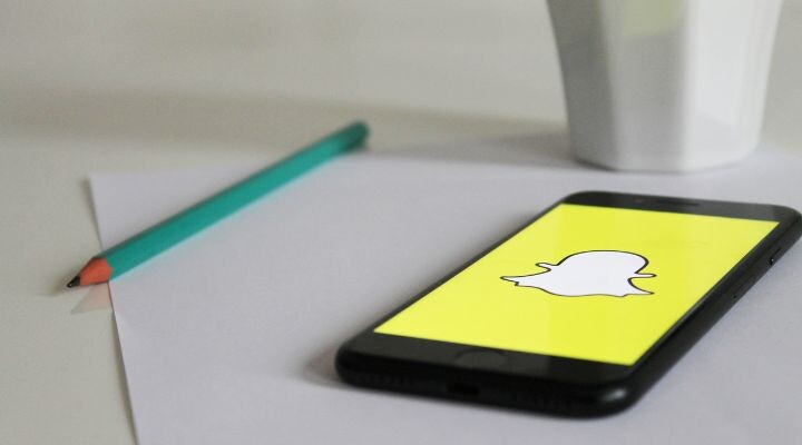 Un teléfono inteligente que muestra el logotipo de Snapchat en una pantalla amarilla, colocado sobre un papel junto a un lápiz verde azulado, puede ilustrar cómo Snapchat como parte de tu estrategia 