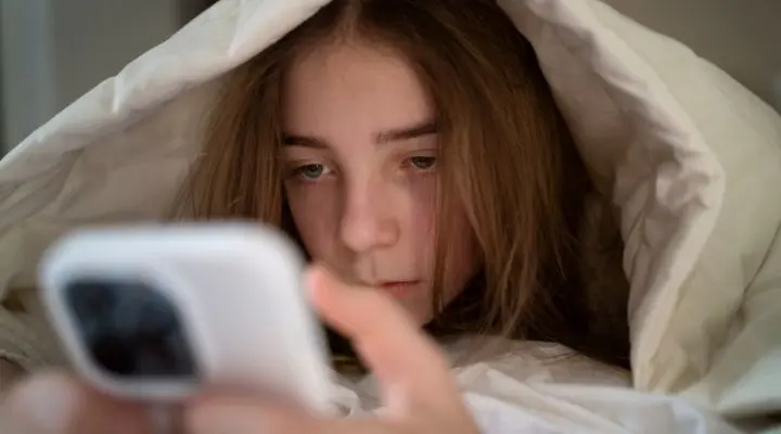 Una persona con cabello largo yace debajo de una manta blanca, mirando su teléfono inteligente, tal vez enviando mensajes de texto mientras sonríe silenciosamente para sí misma a causa de el sexting