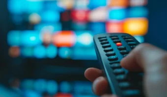 Una mano que sostiene un control remoto de TV, lista para tomar una foto con Izzi, apunta hacia una pantalla de televisión borrosa que muestra múltiples miniaturas coloridas.