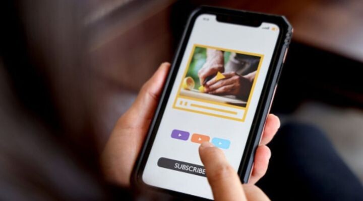 Una persona sostiene un teléfono inteligente que muestra un video de alguien cortando comida. Tres botones coloridos y la opción de suscripción indican el valor de un suscriptor, invitando a los espectadores a participar por correo electrónico para recibir más contenido culinario.