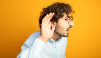 Un hombre con cabello rizado y gafas, con una camisa azul claro, se lleva la mano a la oreja, pareciendo saber si está escuchando, sobre un fondo amarillo sólido.