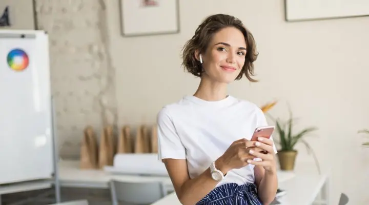 Una mujer que viste una camisa blanca y auriculares inalámbricos sostiene un teléfono inteligente y sonríe mientras está de pie en un espacio de oficina moderno y luminoso, encarnando la confianza y el personal branding