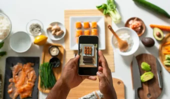 Una persona toma una fotografía de rollos de sushi con un teléfono inteligente, rodeados de varios ingredientes y utensilios en el mostrador de una cocina, perfecto para el marketing gastronómico.