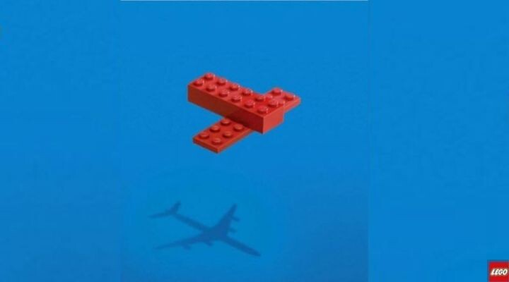 Dos ladrillos LEGO rojos forman la figura de un avión simple sobre un fondo azul, y su sombra proyecta una imagen que recuerda a los aviones reales que suelen verse en los anuncios publicitarios.