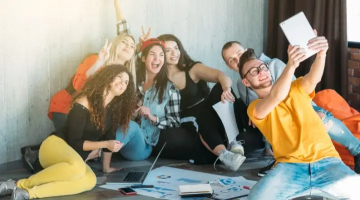 Un grupo de seis millennials están sentados en el suelo, sonriendo y posando para una selfi con una tableta. A su alrededor hay papeles, una computadora portátil y gráficos.