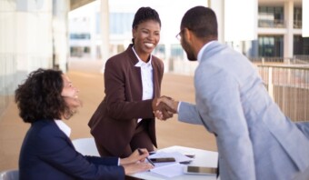 Tres profesionales al aire libre; dos mujeres sentadas, sonriendo, mientras un hombre de pie estrecha la mano de una mujer. Documentos y teléfono sobre la mesa significan una reunión exitosa enfocada en la fidelización de clientes.
