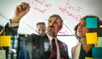 Tres personas vestidas de negocios revisan y escriben notas y gráficos relacionados con la gestión de marketing en un tablero de vidrio cubierto con notas adhesivas de colores y escritura con marcador rojo.