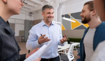 Un hombre con una camisa azul claro habla y gesticula con las manos, pareciendo cacarear el huevo mientras tres colegas escuchan en un entorno de oficina moderno.