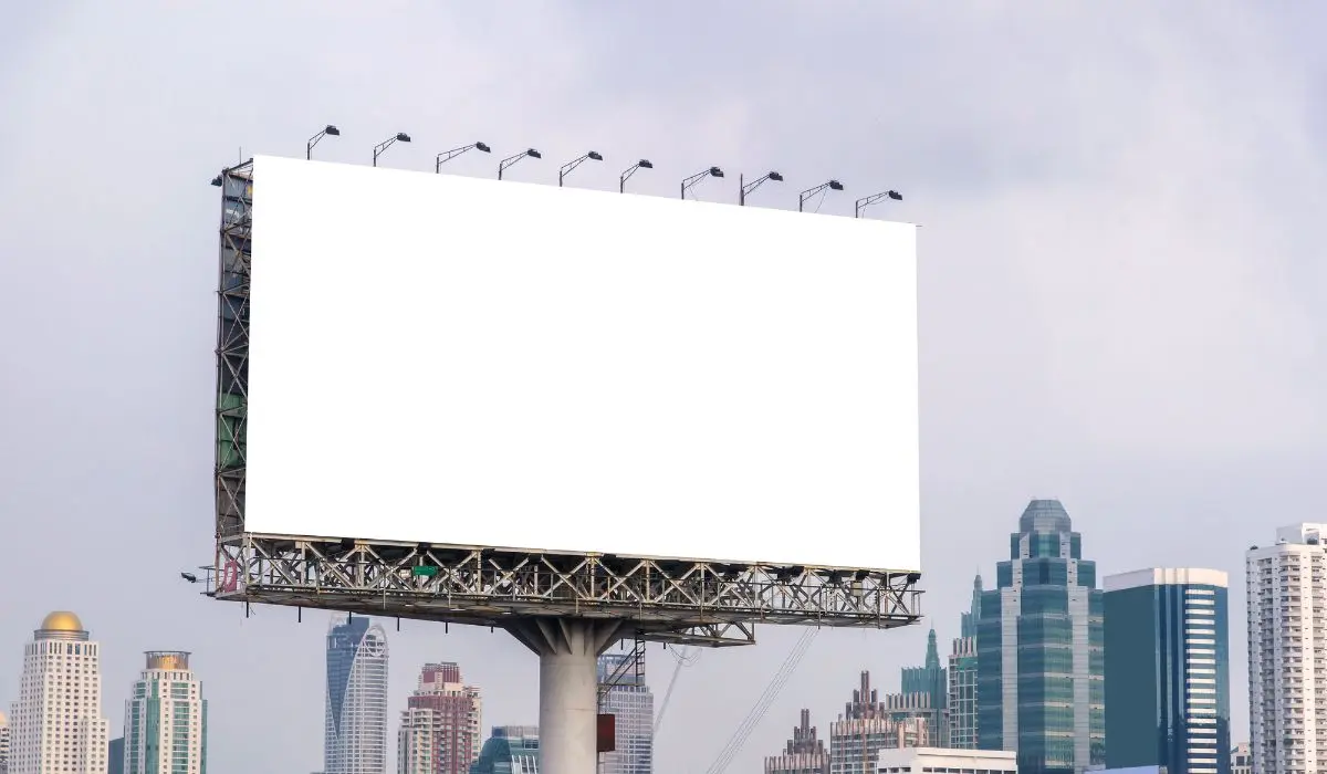 Un gran cartel blanco vacío se encuentra en un poste de metal sobre los edificios de la ciudad contra un cielo nublado, listo para mostrar anuncios creativos para captar la atención de los transeúntes.