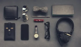 Una disposición plana de artículos necesarios de uso diario, incluidos una billetera, gafas de sol, pajarita, bolígrafo, cuaderno, teléfono, reloj, gafas, auriculares, bolsa de cuero y un pequeño estuche rojo sobre una superficie gris.