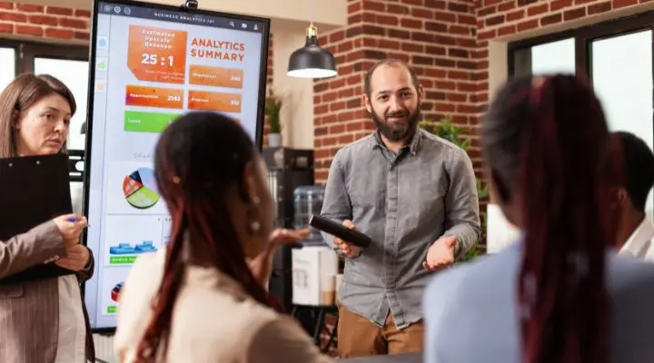 Un hombre presenta un resumen analítico — como si fuera un pitch — a un grupo pequeño en una oficina, mientras una mujer toma notas en un portapapeles.