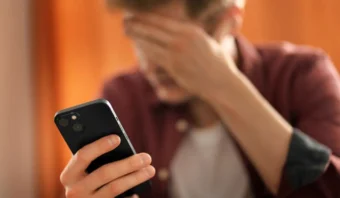 Una persona sostiene un teléfono inteligente en una mano mientras se cubre la cara con la otra, y parece estresada o frustrada mientras lidia con las redes sociales de un fallecido.