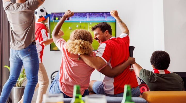 La gente anima y festeja en una sala de estar, envuelta en la emoción de un partido de fútbol en la televisión. Es como una campaña de marketing deportivo improvisada, donde la pasión por el marketing deportivo es contagiosa y une a todos.