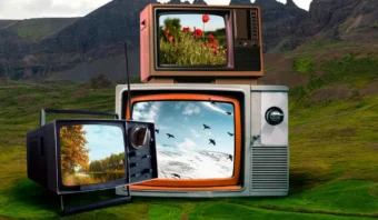 Tres televisores antiguos se apilan sobre el césped, y cada pantalla muestra una escena exterior diferente: amapolas, árboles otoñales y pájaros volando sobre la nieve. Tras ellos se alzan colinas rocosas que invitan a preguntarse: "¿En qué año vive?".