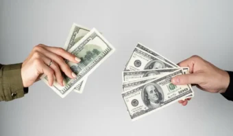 Dos manos intercambiando varios billetes de cien dólares estadounidenses sobre un fondo claro y liso, simbolizando el gasto publicitario.