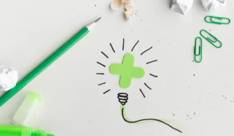 Un signo más verde con líneas garabateadas en forma de bombilla se encuentra sobre una superficie blanca, rodeado de un bolígrafo verde, sujetapapeles, un marcador y papel arrugado: elementos que inspiran a los consumidores a despertar su creatividad.