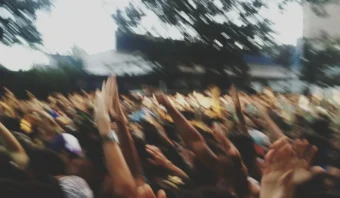 Una multitud grande y borrosa con muchas manos levantadas al aire libre, lo que sugiere movimiento y actividad bajo árboles y edificios en el fondo, captura la energía vibrante del consumidor participando en un evento público animado.