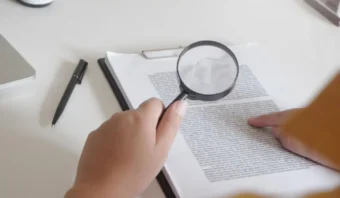 Una persona sostiene una lupa sobre un documento impreso en un portapapeles, examinando detenidamente las letras chiquitas. Cerca, sobre el escritorio blanco, hay un bolígrafo.