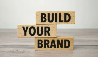 Tres bloques de madera apilados sobre una superficie de madera muestran las palabras "CONSTRUYE TU MARCA" en letras negras en negrita, enfatizando la importancia de desarrollar tu marca personal.