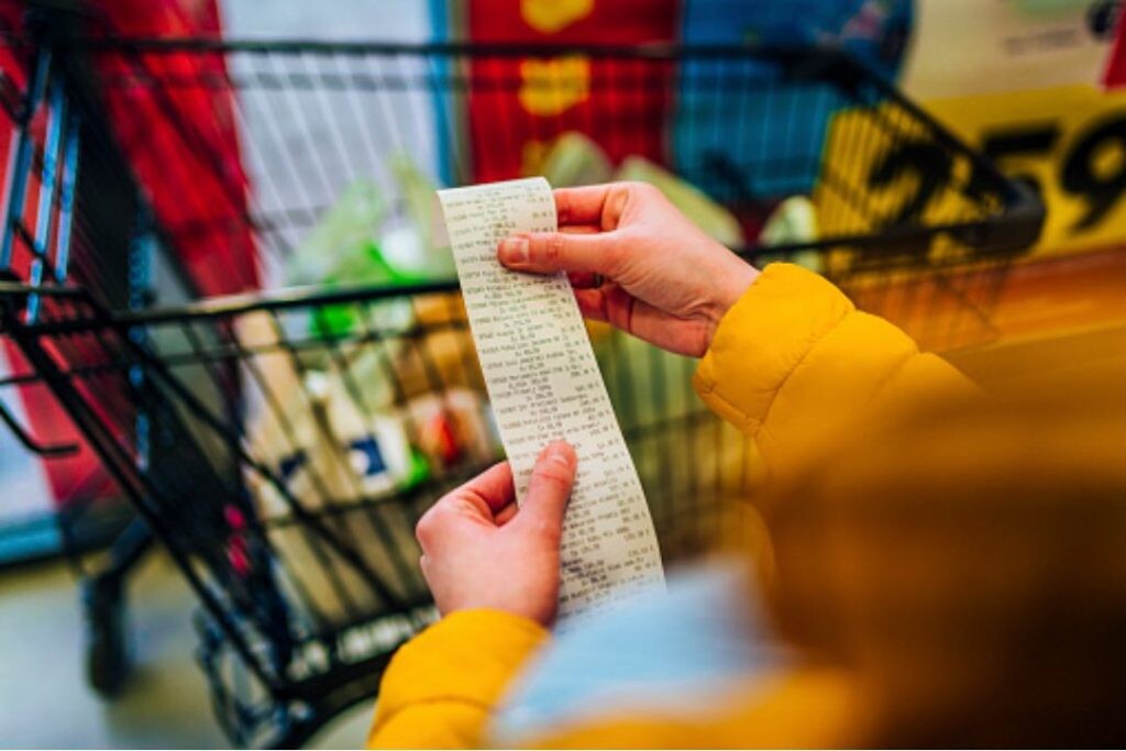 Una persona con una chaqueta amarilla sostiene un recibo largo, de pie orgullosamente junto a un carrito de supermercado repleto de artículos, lo que recuerda a las personas mayores abasteciéndose de lo esencial para su despensa mensual. los seniors 