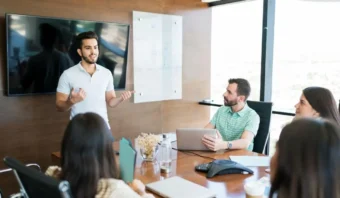 Un hombre se pone de pie y se presenta ante tres colegas sentados alrededor de una mesa de conferencias en una moderna sala de reuniones de una oficina, discutiendo la transformación digital dentro de las organizaciones.