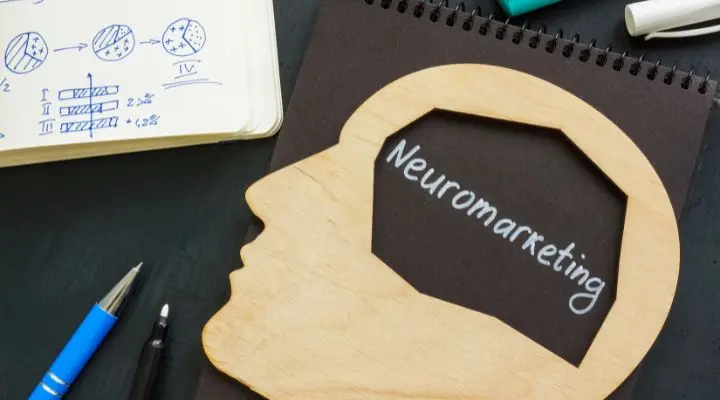 Una silueta de cabeza de madera con la palabra "Neuromarketing" grabado en su interior se encuentra junto a un cuaderno lleno de gráficos circulares vibrantes y un bolígrafo, que ilustran la intersección de la ciencia cognitiva y el marketing estratégico. Seamos serios