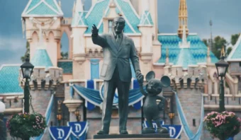 Una estatua de bronce de un hombre tomado de la mano de Mickey Mouse se encuentra frente a un castillo adornado con banderas azules y flores; uno se pregunta si si Walt Disney todavía viviera, estaría orgulloso de este legado mágico.