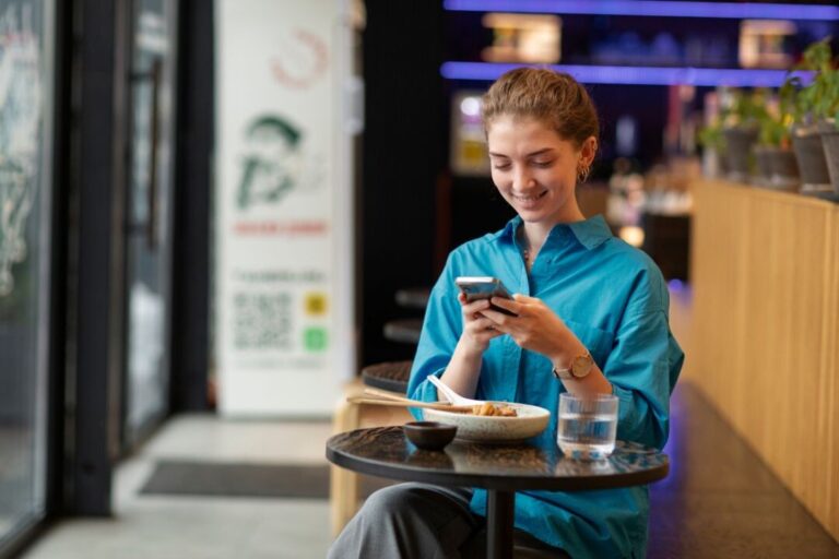 Una mujer está sentada a la mesa de un café comiendo, sonriendo mientras revisa su teléfonopara hacer check in. En el mostrador, a su lado, se ven un vaso de agua y plantas en macetas.