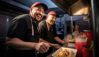 Dos hombres sonrientes con uniformes iguales y sombreros rojos preparan juntos Contenido Fast Food en una concurrida cocina comercial.