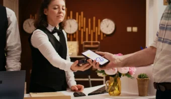 Una recepcionista de hotel uniformada ayuda a un huésped a hacer check in utilizando una terminal de pago para escanear un código QR en el teléfono inteligente del huésped en la recepción.