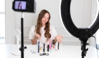 Una mujer se sienta en una mesa con productos de maquillaje, gesticulando con las manos mientras es filmada por una cámara e iluminada por un anillo de luz, una escena común entre los influencers que crean una guía práctica para entusiastas de la belleza.