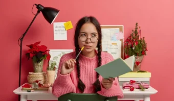 Una mujer joven con cabello trenzado y anteojos se sienta en un escritorio sosteniendo un cuaderno y un bolígrafo, luciendo pensativa (perfecta para ver si te consideras creativo), con plantas, regalos y notas en la pared detrás de ella.