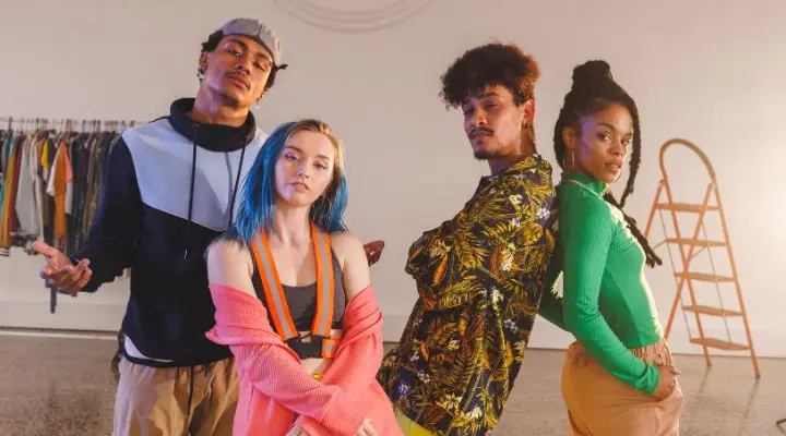 Cuatro jóvenes adultos se encuentran en un interior con ropa elegante y colorida, irradiando confianza y estilo. Al fondo se ven un perchero y una escalera de madera.