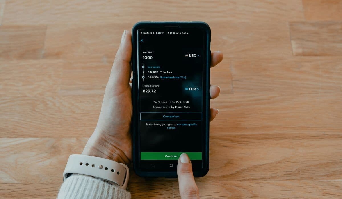 Una persona que sostiene un teléfono inteligente muestra una pantalla de transferencia de dinero internacional con conversión de USD a EUR, mostrando la comodidad del pago móvil sobre una superficie de madera.