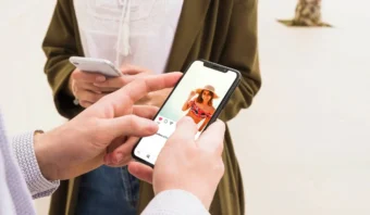Una persona se desplaza por las redes sociales en un teléfono inteligente y ve la foto de una mujer con sombrero (posiblemente una influencer); otra persona sostiene un teléfono en el fondo.