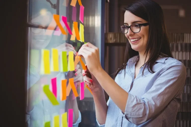 Una mujer que usa gafas, como muchos brand managers, coloca notas adhesivas de colores sobre una superficie de vidrio, sonriendo mientras trabaja en un entorno de oficina moderno.