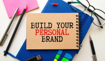 Un bloc de notas con las palabras "CONSTRUYE TU MARCA PERSONAL" se encuentra sobre un escritorio rodeado de un cuaderno azul, gafas, bolígrafos, notas adhesivas, una unidad USB y una chincheta: perfecto para desarrollar tu marca personal.