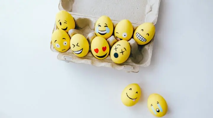Una caja que contiene ocho huevos amarillos pintados con diferentes caras de emoji, incluido un emoji de guiño, con dos huevos más decorados colocados en la superficie blanca cercana.