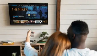 Una pareja está sentada en un sofá, y uno de ellos sostiene un control remoto, disfrutando el entretenimiento de ver un servicio de streaming en la televisión. La pantalla muestra la película "El Caminante".