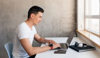 Un hombre con camiseta blanca, representando el estilo millennial de sello, trabaja con una laptop en un escritorio cerca de una ventana. Un teléfono inteligente, un libro, auriculares y audífonos inalámbricos están dispersos por el espacio de trabajo.