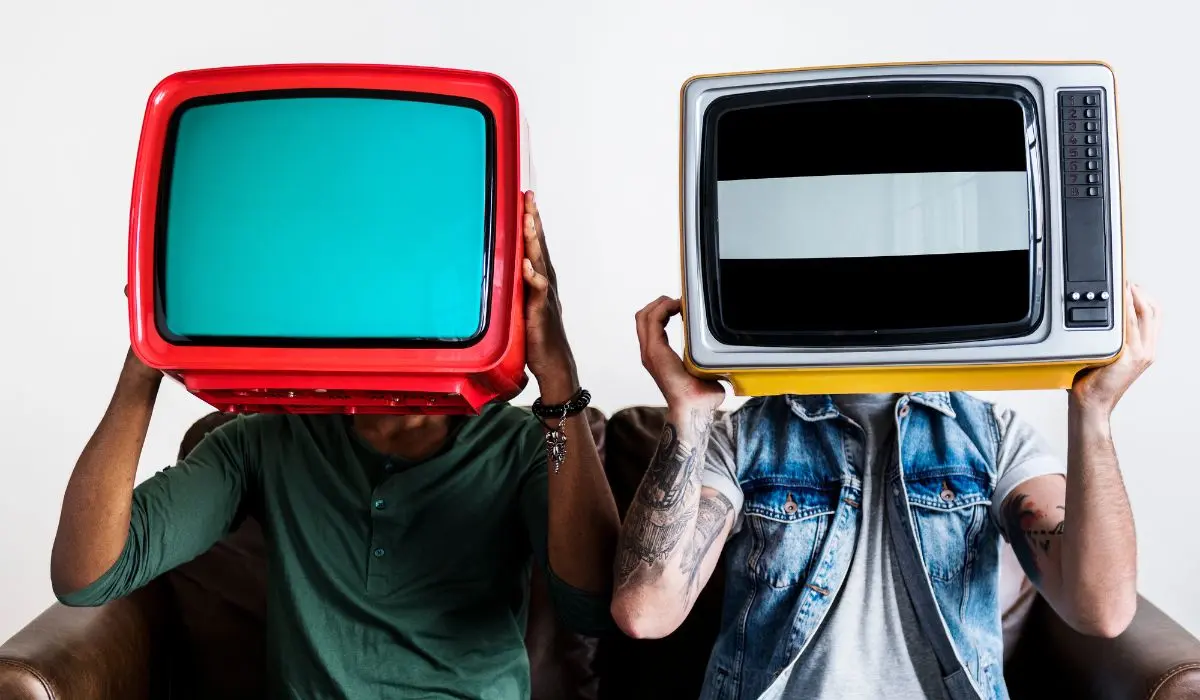 Dos personas se sientan en un sofá con televisores antiguos sobre sus cabezas âuno rojo, otro amarilloâ frente a un fondo blanco liso, encarnando el encanto peculiar de las coincidencias.