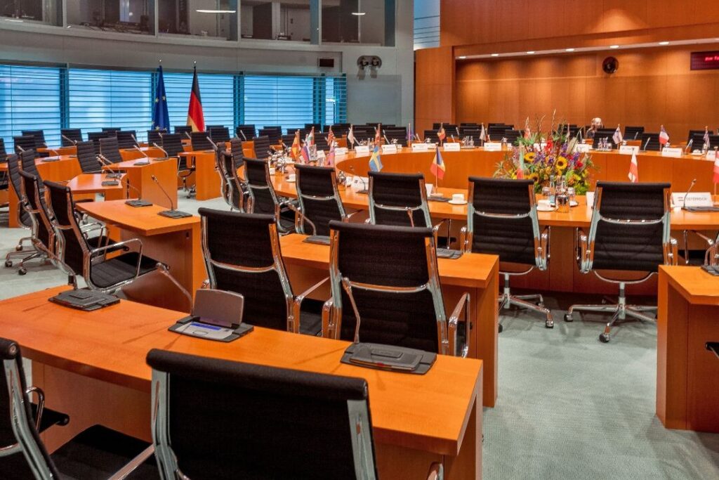 En la sala de conferencias vacía, donde se planifica meticulosamente una campaña de marketing político, mesas circulares de madera se combinan con sillas negras y están flanqueadas por banderas. Un ramo de flores adorna una mesa, mientras que grandes ventanales y un techo alto forman el elegante telón de fondo.