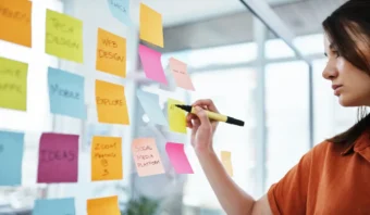 Una persona escribe en una pared de vidrio cubierta con notas adhesivas de colores, organizando tareas e ideas para un plan de marketing de contenidos.