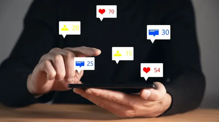Una persona usa un teléfono inteligente con íconos de notificaciones de Redes Sociales para me gusta, comentarios y seguidores flotando sobre la pantalla.