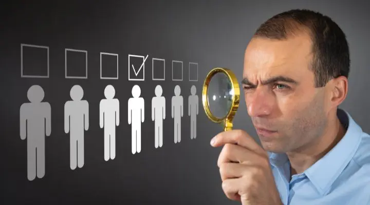 Un hombre sostiene una lupa y mira íconos de personas, uno de ellos marcado con una marca de verificación, que simboliza el proceso para elegir agencia.