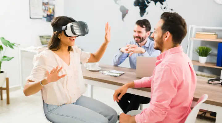 Una mujer que lleva un casco de realidad virtual hace gestos con las manos mientras dos colegas, un hombre y una mujer, sonríen y la observan en un entorno de oficina, experimentando la emoción de la tecnología virtual a lo real.