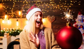 Una mujer con un gorro de Papá Noel sonríe y mira con entusiasmo un gran adorno rojo con una bengala encendida en la parte superior, rodeado de luces navideñas festivas, celebrando las promesas cumplidas en esta alegre temporada.