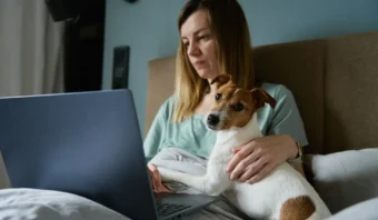 Una mujer se sienta en la cama usando una computadora portátil con un pequeño perro en su regazo, ambos mirando la pantalla de la computadora portátil, desafiando la noción de que tales escenas son sexistas.