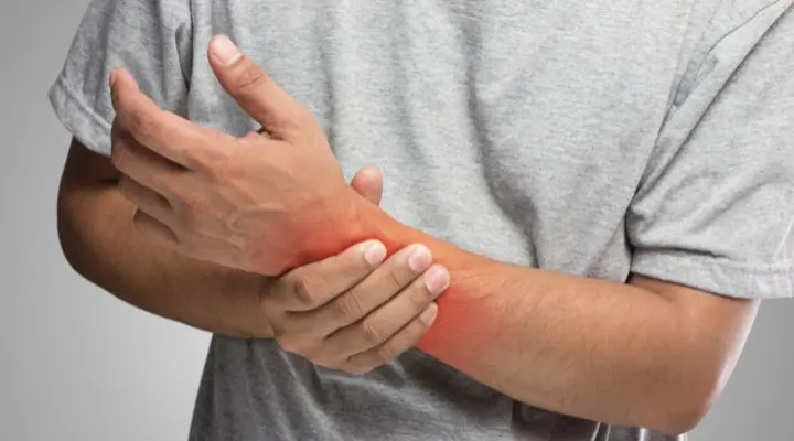 Una persona que viste una camiseta gris sostiene su muñeca izquierda con su mano derecha; el área de la muñeca está resaltada en rojo, lo que ilustra un punto de dolor o malestar o mejor conocido como pain point.